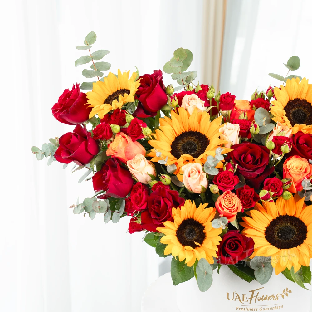 A box arrangement of 6 sunflowers, 12 red roses, 3 red spray roses, 2 orange spray roses and 2 pink spray roses with green fillers.