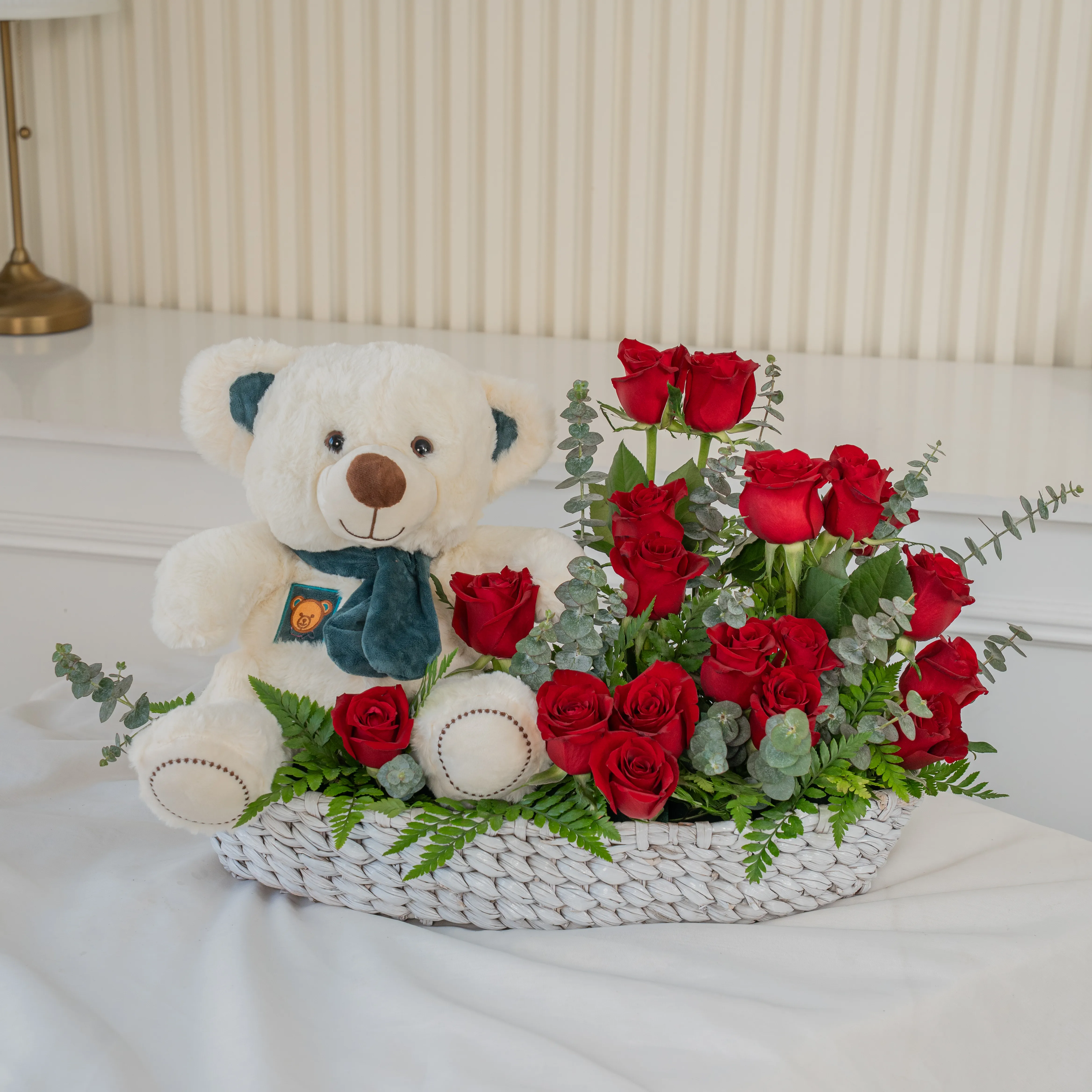 Basket arrangement of 20 red roses and small white teddy bear with eucalyptus leaf and green fillers.