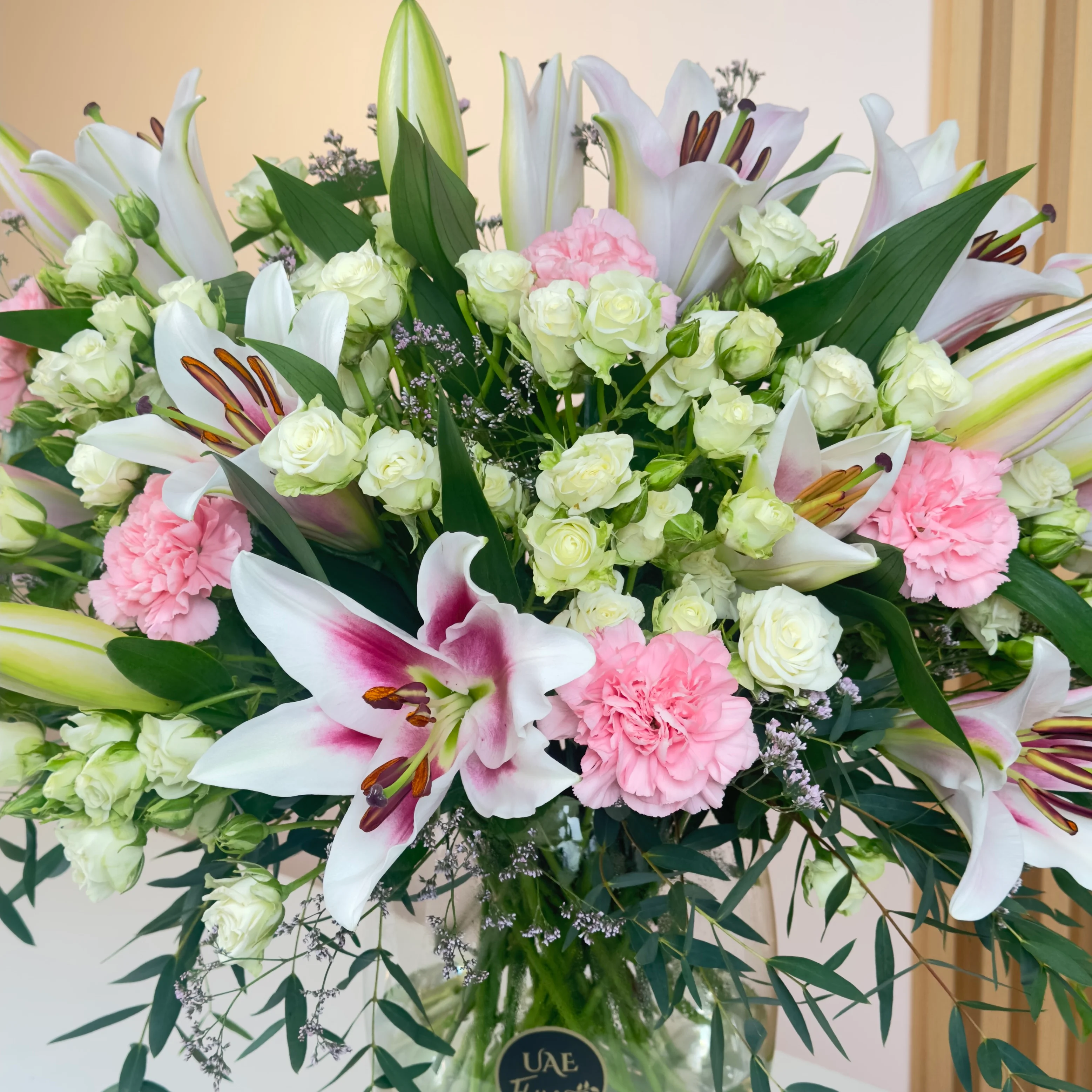 An arrangement of 4 pink lilies, 10 pink carnations, and 10 white spray roses with green fillers in a glass vase