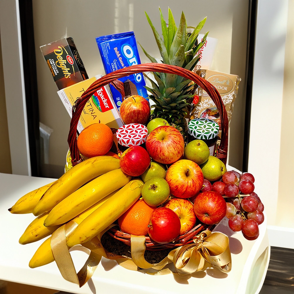 A large basket containing fresh pineapple, apples, pears, bananas, and red grapes, along with a premium selection of biscuits, chocolates, and crackers, adorned with a large bright bow