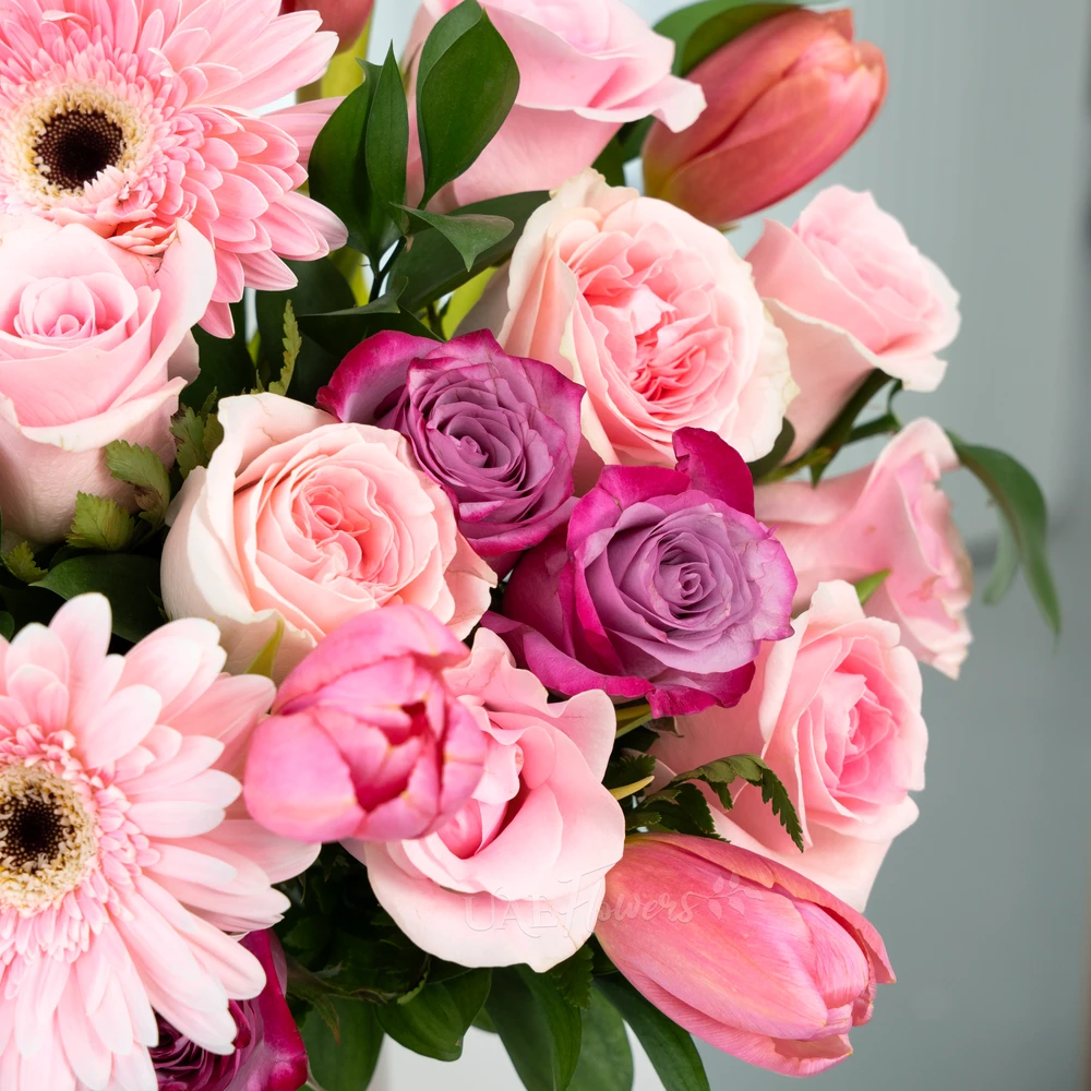 A vase arrangement of 3 pink gerberas, 7 purple roses, 15 pink roses, 10 pink tulips with green fillers and multi colored easter eggs.