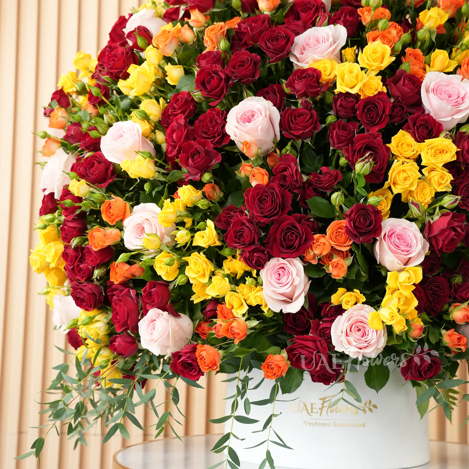 A half-round floral arrangement featuring 30 pink roses and 50 red, orange, and yellow spray roses, along with green fillers and eucalyptus leaves, presented in a white round box