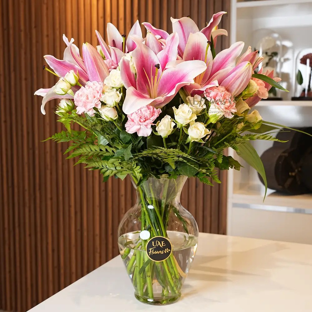 Glass vase with 4 pink lilies, 6 pink carnations, 8 white spray roses, and green fillers, elegantly arranged.