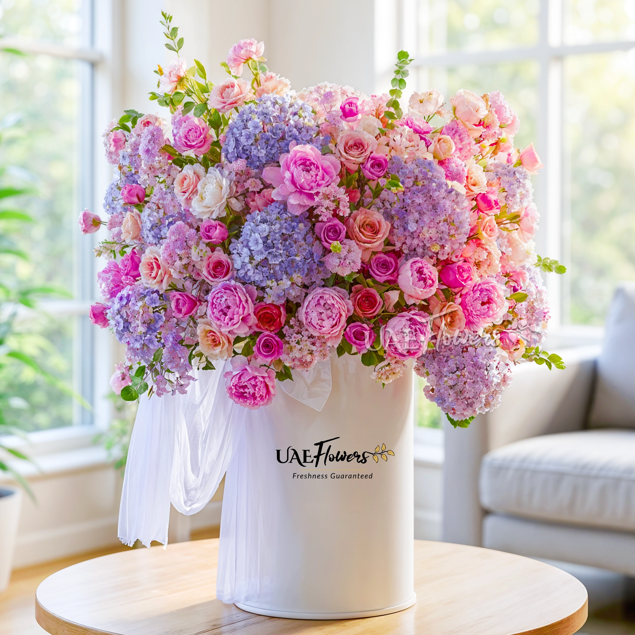 An arrangement of blue hydrangeas, pink peonies, purple roses, purple lisianthus, peach roses and purple chrysanthemums with green fillers and blue net in a tall round box.