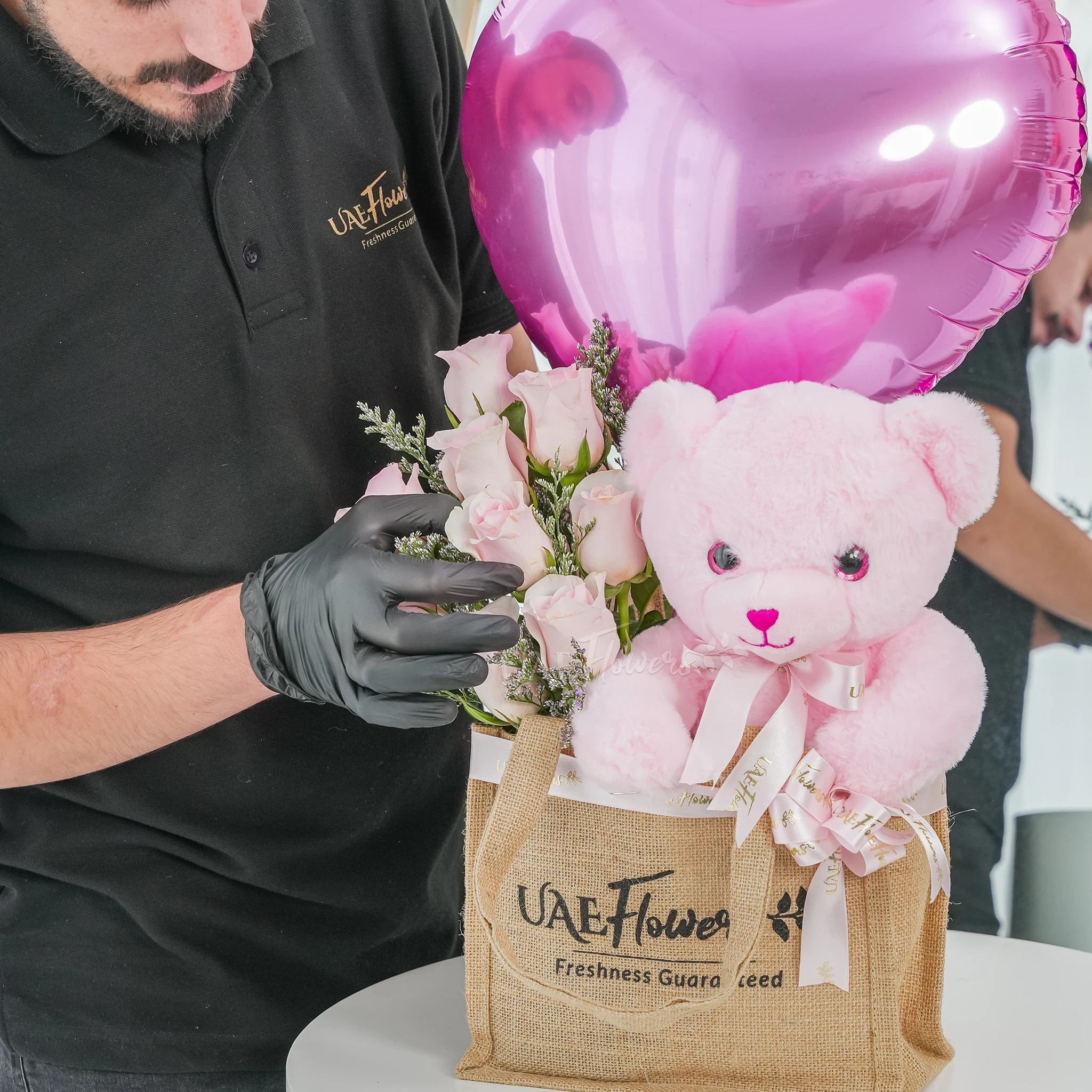 An arrangement of 7 pink roses, small size teddy bear and pink heart shaped balloons in a jute bag